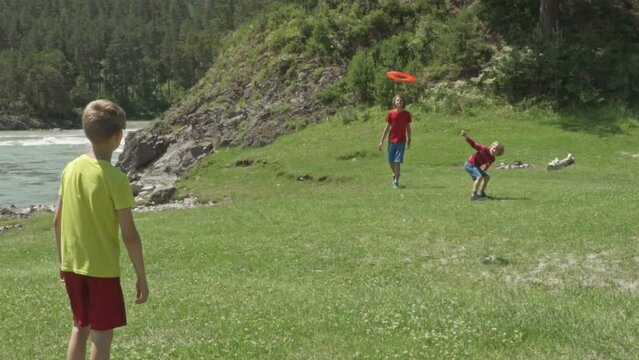 Cute little children playing with frisbee outdoors on sunny day. Against the backdrop of mountains, on the coast of a mountain river. Slow motion. Young Boys throwing a red frisbee disk. 