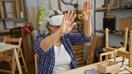A middle-aged woman in a plaid shirt uses vr technology in a woodworking workshop.