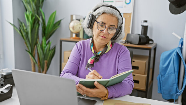 A focused mature woman with grey hair works in an office, writing notes while wearing headphones.