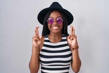 Young african american with braids wearing hat and sunglasses gesturing finger crossed smiling with hope and eyes closed. luck and superstitious concept.