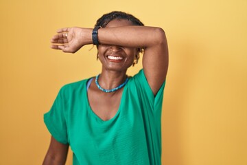 African woman with dreadlocks standing over yellow background covering eyes with arm smiling...