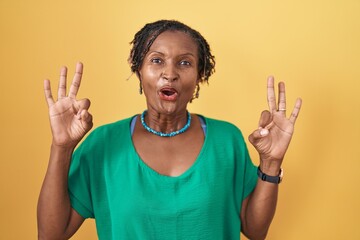 African woman with dreadlocks standing over yellow background looking surprised and shocked doing ok approval symbol with fingers. crazy expression