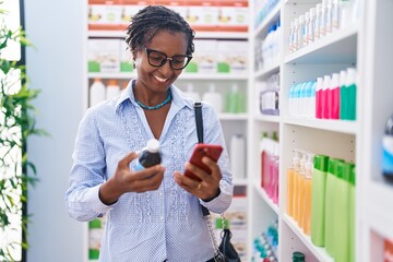 Middle age african american woman customer using smartphone holding medicine bottle at pharmacy