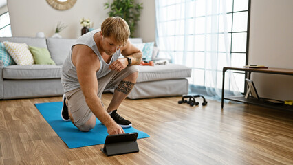 A young blond man with a beard using a tablet while exercising on a yoga mat in a modern living...