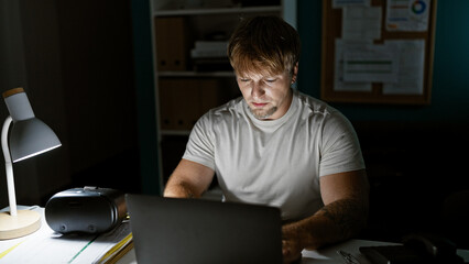 Focused young blond man with beard working late on laptop in dim office room.