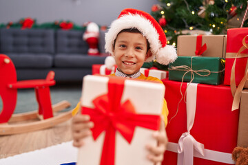 Adorable hispanic boy holding christmas gift sitting on floor at home