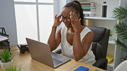 A professional african-american woman feeling stressed in a modern office setting indoors.