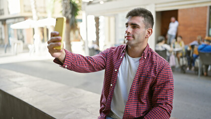 Cool, attractive hispanic guy making self portrait with phone - young man sitting on city bench, capturing his lifestyle through selfie under sunlight outdoors