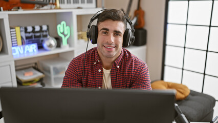 Handsome young hispanic man confidently smiling while sitting at the music studio, headphones on,...