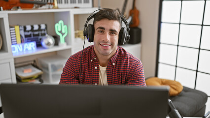 Handsome young hispanic man confidently smiling while sitting at the music studio, headphones on, engrossed in creating tunes, embodying the soul of an attractive musician at work