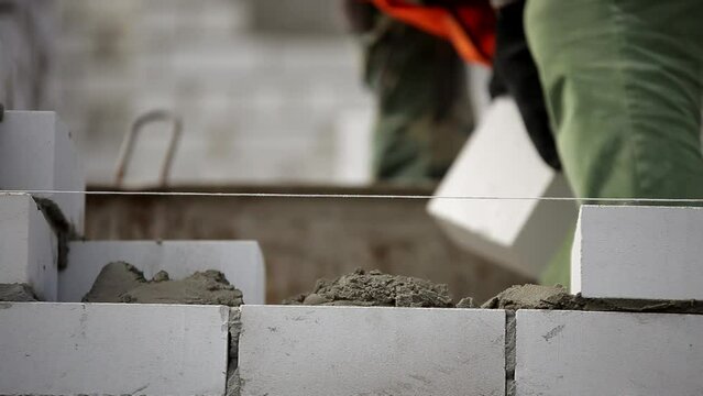 builder applies cement mortar with trowel, then lays white bricks close up slow motion camera. A bricklayer works on the construction of a new high-rise building on a sunny summer day.