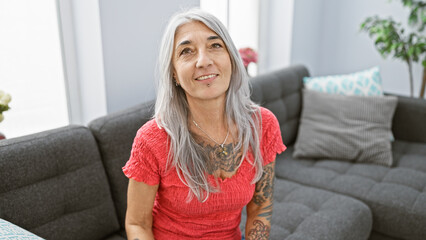 Confident middle age grey-haired woman enjoys a relaxing moment, cheerfully smiling while sitting comfortably on her cozy living room sofa at home
