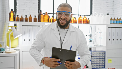 Smiling african american man with beard wearing lab coat and safety goggles holding clipboard in a...