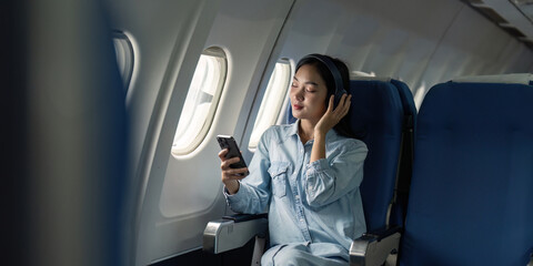 Asian woman traveler in airplane wearing headset listening music from mobile phone going on a trip vacation travel concept