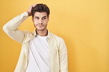 Young hispanic man standing over yellow background confuse and wondering about question. uncertain with doubt, thinking with hand on head. pensive concept.