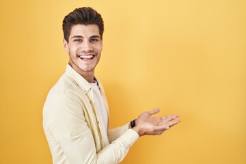 Young hispanic man standing over yellow background pointing aside with hands open palms showing copy space, presenting advertisement smiling excited happy