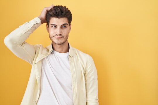 Young hispanic man standing over yellow background confuse and wondering about question. uncertain with doubt, thinking with hand on head. pensive concept.