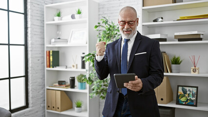 A senior man in a suit celebrates a success with a fist pump while holding a tablet in a modern office.