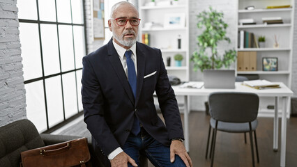 Confident mature businessman with beard in modern office, wearing glasses and blue suit.