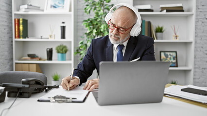 A mature businessman in headphones works at his laptop in a modern office, writing notes.