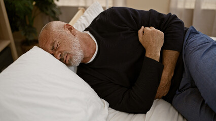 A mature hispanic man with gray hair and a beard clutches his abdomen in pain while lying in a bedroom. - obrazy, fototapety, plakaty