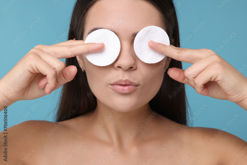 Wall mural Young woman covering her eyes with cotton pads on light blue background, closeup