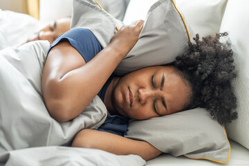 black African American woman covers her ears with a pillow, can't sleep, Asian husband's snoring...