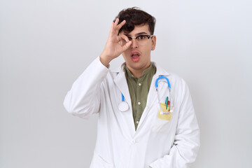 Young non binary man wearing doctor uniform and stethoscope doing ok gesture shocked with surprised face, eye looking through fingers. unbelieving expression.