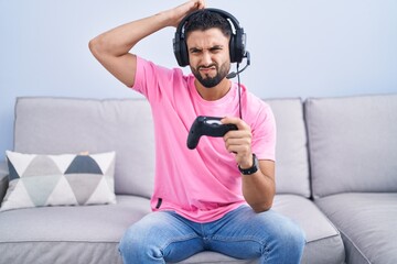 Hispanic young man playing video game holding controller sitting on the sofa confuse and wondering...
