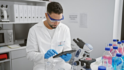 Bearded man in lab coat using tablet in a modern laboratory with microscope and specimens.