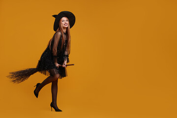 Happy young woman in scary witch costume with broom posing on orange background, space for text....