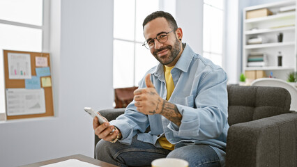 Handsome hispanic man with beard giving thumbs-up in modern office setting holding smartphone