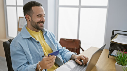 Handsome hispanic man with a beard shopping online using a credit card and laptop in a modern...