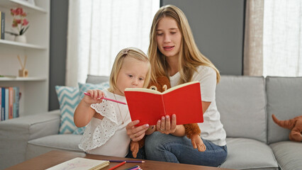 Serious caucasian mother and little daughter enjoy reading a book together, relaxing at home on the sofa surrounded by toys