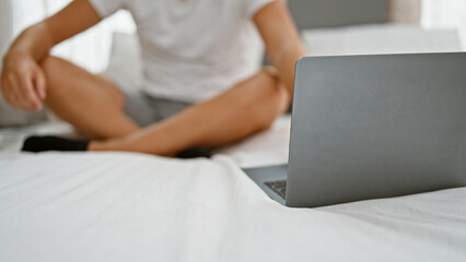 A casual young man sitting on a bed, with a laptop open in front of him in a bright bedroom