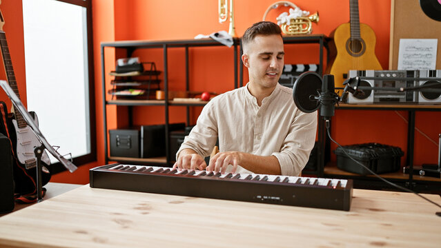 A young hispanic man plays keyboard in an orange music studio full of instruments.