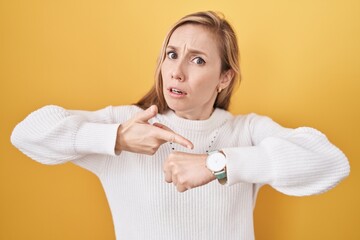 Young caucasian woman wearing white sweater over yellow background in hurry pointing to watch time,...