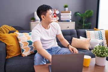 Young asian man using laptop at home sitting on the sofa looking to side, relax profile pose with...