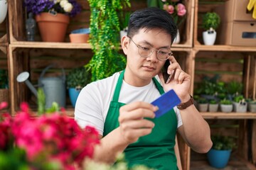 Young chinese man florist talking on smartphone holding credit card at flower shop