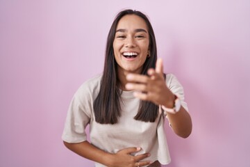 Young hispanic woman standing over pink background laughing at you, pointing finger to the camera with hand over body, shame expression