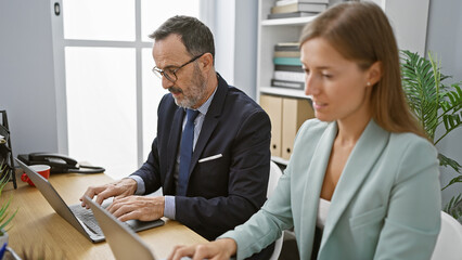 Two serious-faced business workers engrossed in a project, sitting together at the office desk, working on a laptop, embodying professional success
