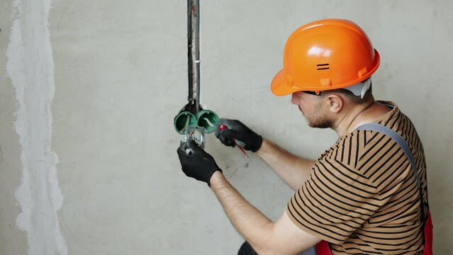Electrician male in uniform, protective gloves and helmet checks presence of electrical voltage in socket phase uses electrical tester screwdriver. Examining wires in outlet by voltage detector indoor