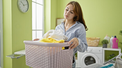 Naklejka na ściany i meble Young woman holding basket with clothes standing with sad expression at laundry room