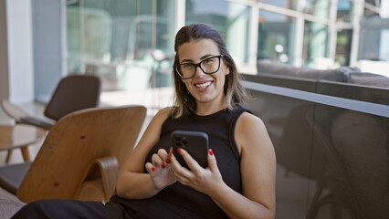 Radiant hispanic woman, all smiles, busily messaging on her smartphone while fashionably lounging...