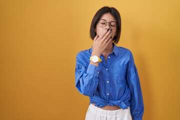 Young girl standing over yellow background bored yawning tired covering mouth with hand. restless and sleepiness.