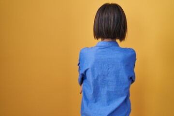 Young girl standing over yellow background standing backwards looking away with crossed arms