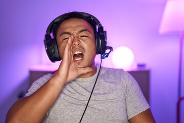 Chinese young man playing video games wearing headphones clueless and confused with open arms, no...