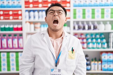 Chinese young man working at pharmacy drugstore angry and mad screaming frustrated and furious,...
