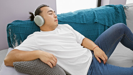 Handsome young chinese man, chilled out while tuned into music on his headphones, sinks into his...