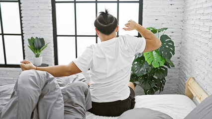 Cozy morning as a handsome young chinese man wakes up, stretches arms back in bed while sitting up,...